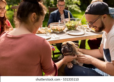 Leisure, Holidays, Eating, People And Food Concept - Happy Friends With French Bulldog Dog Having Dinner At Summer Garden Party