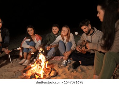 leisure and people concept - group of smiling friends sitting at camp fire on beach and roasting marshmallow at night - Powered by Shutterstock