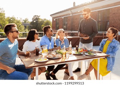 Leisure And People Concept - Happy Party Host Offering Meat To His Friends At Barbecue Party On Rooftop In Summer