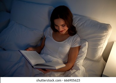 Leisure And People Concept - Young Woman Reading Book In Bed At Night Home