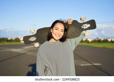 Leisure and people. Happy asian woman standing with longboard, cruising on an empty road in countryside. Skater girl holds her skateboard and smiles at camera.