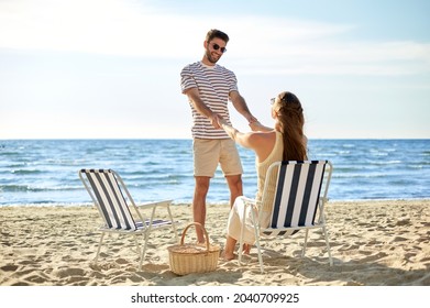 leisure, relationships and people concept - happy couple with picnic basket sitting in folding chairs and holding hands on summer beach