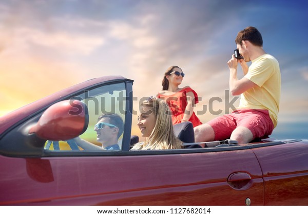 leisure, road trip, travel,
summer holidays and people concept - happy friends driving in
convertible car and taking picture by film camera over sky
background