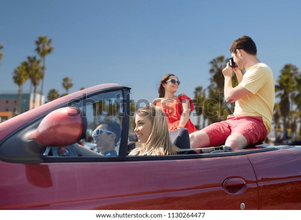 leisure, road trip, travel, summer holidays\
and people concept - happy friends driving in convertible car and\
taking picture by film camera over venice beach background in\
california