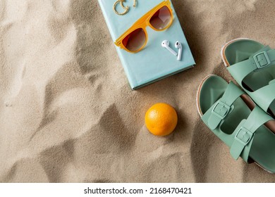 leisure and summer holidays concept - slippers, orange, earbuds, book and sunglasses on beach sand