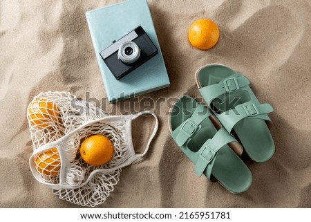 leisure and summer holidays concept - slippers, string bag of oranges, film camera and book on beach sand