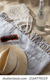 Leisure And Summer Holidays Concept - String Bag, Sunglasses, Glass Bottle Of Water, Straw Hat And Beach Blanket On Sand