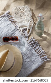 Leisure And Summer Holidays Concept - String Bag, Sunglasses, Glass Bottle Of Water, Straw Hat And Beach Blanket On Sand