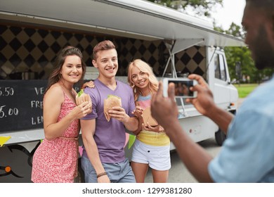 Leisure, Technology And People Concept - Young Man Taking Picture Of His Happy Friends Eating Hamburgers And Wok At Food Truck