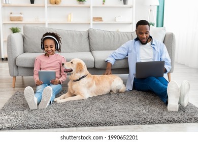 Leisure Time. African American girl sitting on rug floor carpet with dog and holding digital tablet, wearing headphones, man using computer and working at home office browsing internet, patting dog