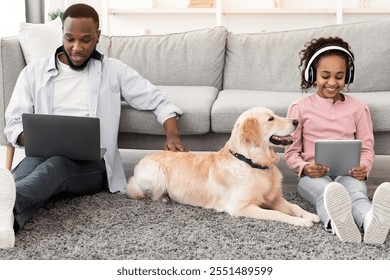 Leisure Time. African American girl sitting on rug floor carpet with dog and holding digital tablet, wearing headphones, man using computer and working at home office browsing internet, patting dog - Powered by Shutterstock