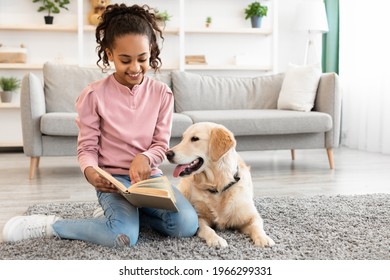 Leisure Time Concept. Portrait Of Cheerful Black Girl Reading Paper Book And Showing Page To Dog, Sitting On The Floor Carpet With Golden Retiever In Living Room Or Bedroom At Home, Having Fun