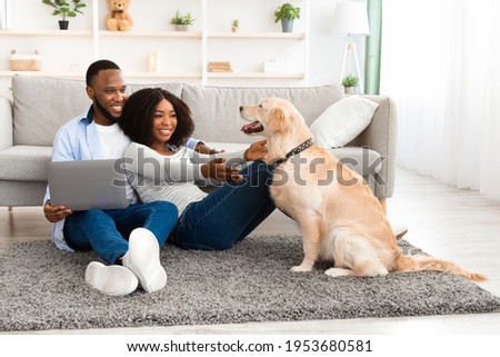 Leisure Time. Portrait of loving black family sitting on floor carpet, man higging lady and using laptop, woman playing with labrador. Married couple spending time together at home in living room