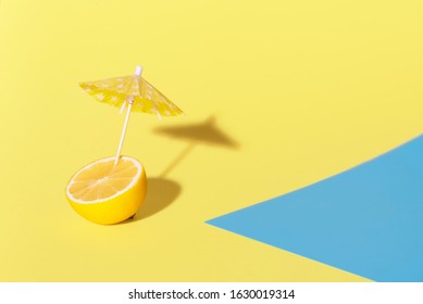 Lemon With A Cocktail Umbrella In It, In Sunlight, On A Yellow Background Near A Blue Patch. Summer Drink Over The Pool Concept. Citrus In Summer Sun.