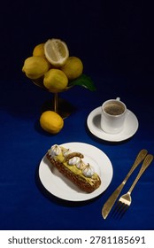 Lemon eclair on a plate next to a cup of coffee and a vase filled with fresh lemons on a blue table surface.
