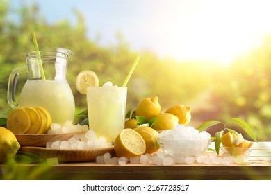 Lemonade with ice in a pitcher and glass on a wooden table with fruit and crushed ice outside with a lemon field in the background on a sunny day. Front view. Horizontal composition. - Shutterstock ID 2167723517