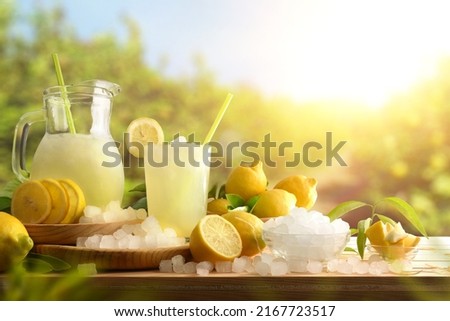 Lemonade with ice in a pitcher and glass on a wooden table with fruit and crushed ice outside with a lemon field in the background on a sunny day. Front view. Horizontal composition.