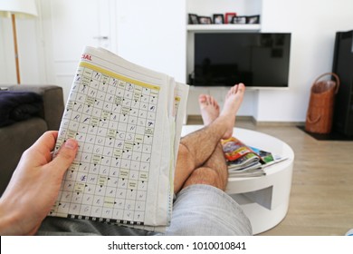 LENDELEDE,BELGIUM-APRIL 7TH 2017:Hand holding a filled in crossword puzzle in the newspaper at home.The person is laying with his feet on the table, with a TV in the background. Editorial illustrative