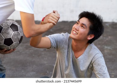 Lending A Helping Hand. Asian Boy Helping His Friend Up From The Ground During A Soccer Game.