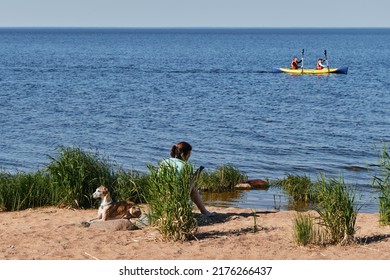 Leningrad Region, Russia - June 2022. Female Owner With Whippet Dog On Sandy Riverbank In Summer. Man And Woman In Life Jackets Row Oars Sitting In Kayak On Clear Lake Ladoga.