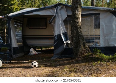 Leningrad Region, Russia - June 2022. Large Trailer For Family Camping In Woods. Nearby There Is Canopy, Camping Furniture And Black And White Soccer Ball. There Are No People.