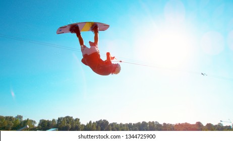 LENS FLARE: Athletic Young Male Wakeboarder Does A Backflip Across The Blue Sky. Spectacular Shot Of Extreme Athlete Doing A Cool Trick While Wakesurfing In A Water Park On A Perfect Summer Day.