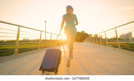 LENS FLARE: Blonde Haired Tourist Girl In Heels Sprints With Her Heavy Luggage To Catch Her Flight. Cinematic Shot Of An Unrecognizable Young Woman Rushing To The Airport On A Sunny Summer Evening.