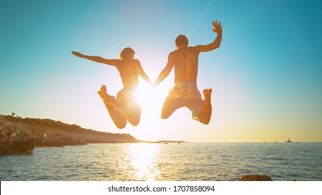 LENS FLARE: Carefree tourists hold hands while jumping into the refreshing sea at golden sunset. Young and energetic traveler couple holds hands while diving into the sea on a sunny summer evening. - Powered by Shutterstock