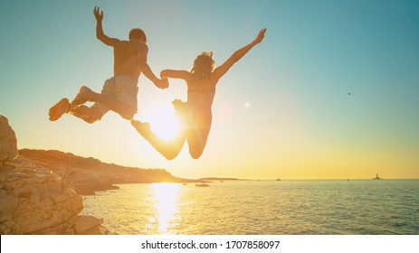 LENS FLARE: Carefree tourists hold hands while jumping into sea at sunset. Cheerful young woman holds her athletic boyfriend's hand as they jump off a cliff and into the cool sea at golden sunset.
