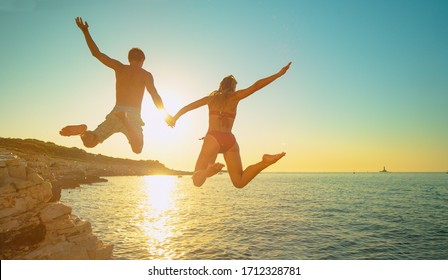 LENS FLARE: Cheerful Caucasian woman holds her boyfriend's hand as they jump off a cliff and into the sea at golden sunset. Fit and carefree tourists hold hands while jumping into sea at sunset. - Powered by Shutterstock