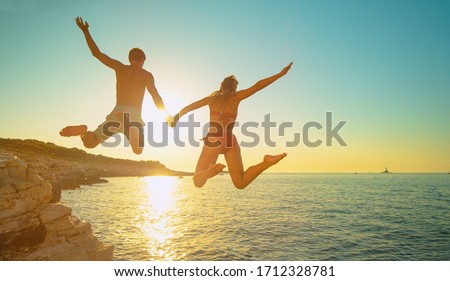 LENS FLARE: Cheerful Caucasian woman holds her boyfriend's hand as they jump off a cliff and into the sea at golden sunset. Fit and carefree tourists hold hands while jumping into sea at sunset.