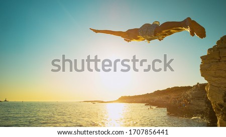 LENS FLARE, CLOSE UP: Athletic man dives off a cliff and head-first into the ocean at sunset. Fit male tourist on vacation jumps of a rocky ledge and dives into the refreshing sea water in Croatia.