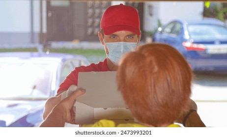 LENS FLARE, CLOSE UP, DOF: Elderly woman opens door for young male courier wearing a protective mask handing her two boxes of pizza. Handsome man delivers pizzas to lady during covid-19 pandemic. - Powered by Shutterstock