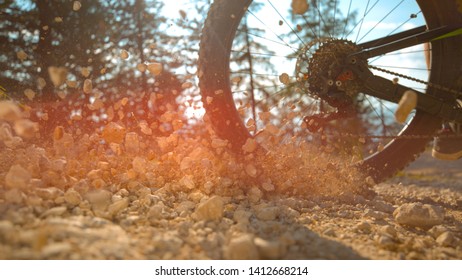 LENS FLARE, CLOSE UP, LOW ANGLE: Unrecognizable athletic man brakes while riding bike down a gravel track. Cinematic shot of rocks flying as mountain cyclist rides down hill on a sunny summer day.