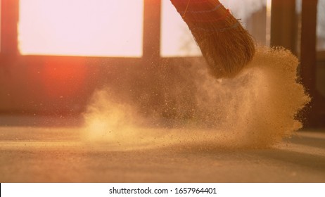 LENS FLARE, CLOSE UP, LOW ANGLE: Dust gets swept up into air as an unrecognizable person cleans the construction site floor. Contractor sweeps the ground of a construction site with a straw broom