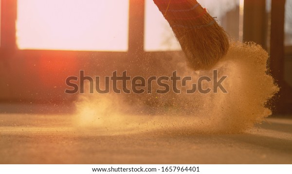 LENS FLARE, CLOSE UP, LOW ANGLE: Dust gets swept up\
into air as an unrecognizable person cleans the construction site\
floor. Contractor sweeps the ground of a construction site with a\
straw broom