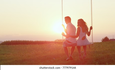 LENS FLARE: Delighted smiling couple swaying on a swing during a romantic sunset. Young girl and boy affectionately looking back and laughing on swing overlooking a green field in the summertime.
