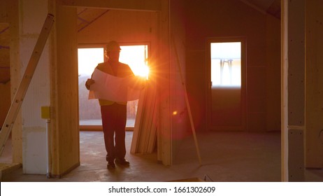 LENS FLARE, DOF: Sunset illuminates the hallways as static engineer walks around the cross-laminated timber house with the floor plans in hand. Architect walks around a CLT house under construction