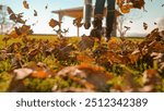 LENS FLARE, DOF: Woman clearing autumn leaves from the garden with an electric and portable leaf blower. Dry brown foliage flies across sunlit grass towards the camera. Garden cleanup in fall season.