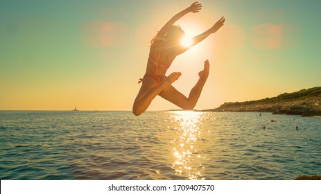 LENS FLARE: Energetic Girl Jumps Off A Cliff And Kicks Legs Back While Diving Into The Ocean On A Sunny Summer Evening. Carefree Young Woman Does An Attractive Dive Into The Refreshing Sea At Sunrise.