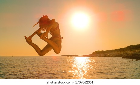 LENS FLARE: Golden Morning Sunbeams Shine On Young Agile Woman Cliff Diving At Sunrise. Unrecognizable Woman Arches Her Back And Kicks Her Legs Back While Jumping Into The Sea On A Sunny Evening