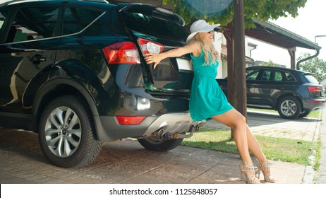 LENS FLARE: Happy Woman In Green Dress Leaning On The Lid Of Her Black Car To Close The Stuffed Trunk. Cheerful Blonde Haired Girl In Heels Pushing The Door Of Her Overpacked Boot To Close It Shut.