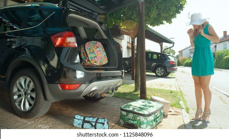 LENS FLARE: Lighthearted Caucasian Woman Fails To Close Her Car And Drops Her Luggage All Over The Driveway In Front Of Her Home In The Suburban Neighborhood. Girl Packs Too Many Bags Into Her Car.