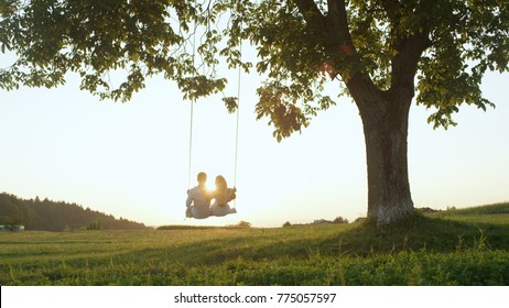 LENS FLARE SILHOUETTE: Setting Sun Shining Over Young Couple's Shoulders On Rope Swing. Young Adults On Romantic Summer Date, Leaning Back Embraced On Swing Under Tree And Watching The Golden Sunset