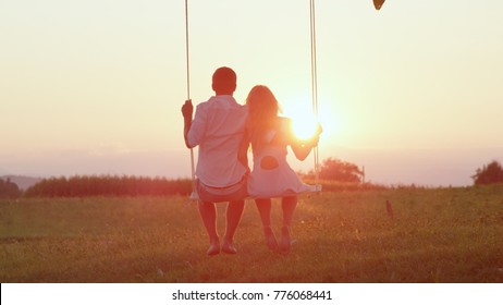 LENS FLARE SILHOUETTE: Unrecognizable couple in love swaying on tree swing at golden summer sunset. Happy man and woman swinging under a tree in sunny evening. Romantic boy and girl embracing on swing