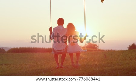 LENS FLARE SILHOUETTE: Unrecognizable couple in love swaying on tree swing at golden summer sunset. Happy man and woman swinging under a tree in sunny evening. Romantic boy and girl embracing on swing