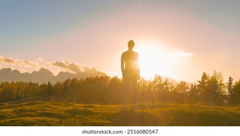 LENS FLARE, SILHOUETTE: Woman stands still, gazing toward the setting autumn sun in a vast mountain landscape. Moment of peaceful reflection and connection with nature in breathtaking alpine scenery. - Powered by Shutterstock