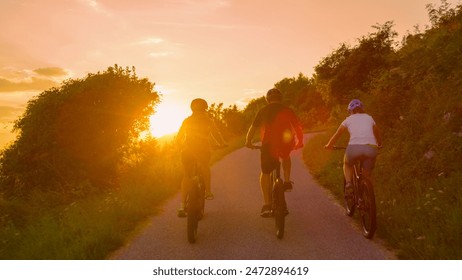 LENS FLARE: Three fit friends enjoy a scenic bicycle journey around the picturesque countryside on an idyllic summer evening. Young travelers explore the rural landscape on electric bikes at sunset. - Powered by Shutterstock