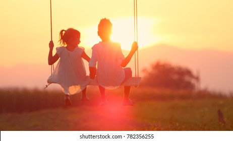 LENSE FLARE Adorable siblings hold hands swaying on late summer evening. Little brother and sister swinging on wood swing in meadow gazing at warm golden sunset. Young love blooming at gold sundown - Powered by Shutterstock