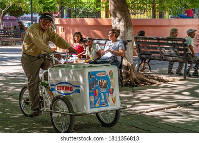 Leon, Nicaragua - November 27, 2008: ice cream vendor on tricycle selling Eskimo brand, while 3 ladies sit on bench in park. Green foliage.
